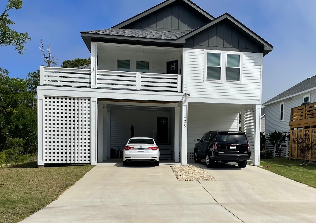 A two-story house is viewed from the front, featuring a spacious driveway with parking for two vehicles. The exterior showcases a combination of white siding and dark gray accents. A covered porch is present above, complemented by a white railing. Lush greenery surrounds the property.