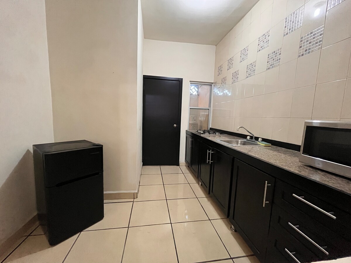 A functional kitchen space includes dark cabinetry and a granite countertop. Essential appliances such as a refrigerator and microwave are present. The tiled floor reflects light, while a window allows natural illumination. The layout is designed for efficient use, enhancing convenience.