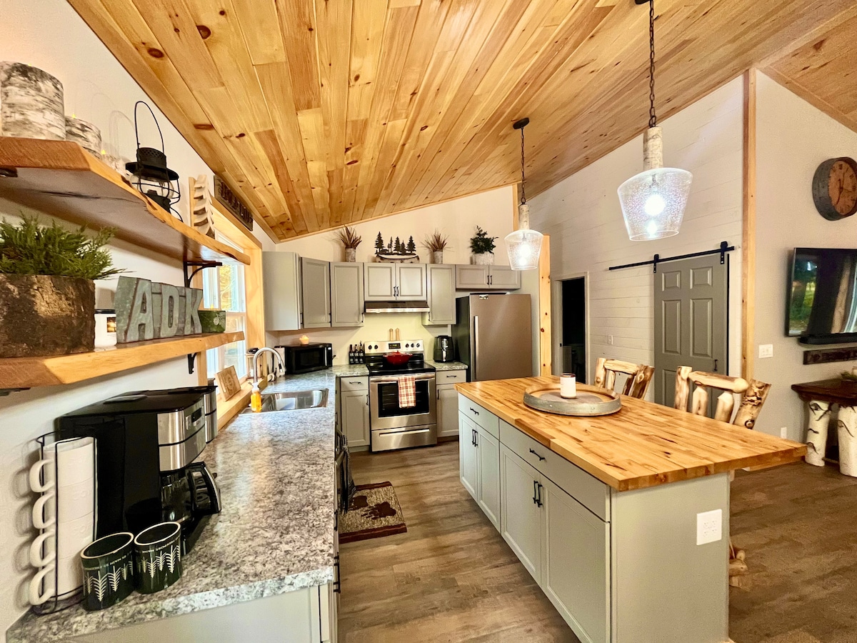 A modern kitchen features wooden beam ceilings and sleek cabinetry. Stainless steel appliances are complemented by a spacious island with a wood countertop. Open shelving displays decorative items, and a cozy dining area is visible, enhancing the inviting layout.
