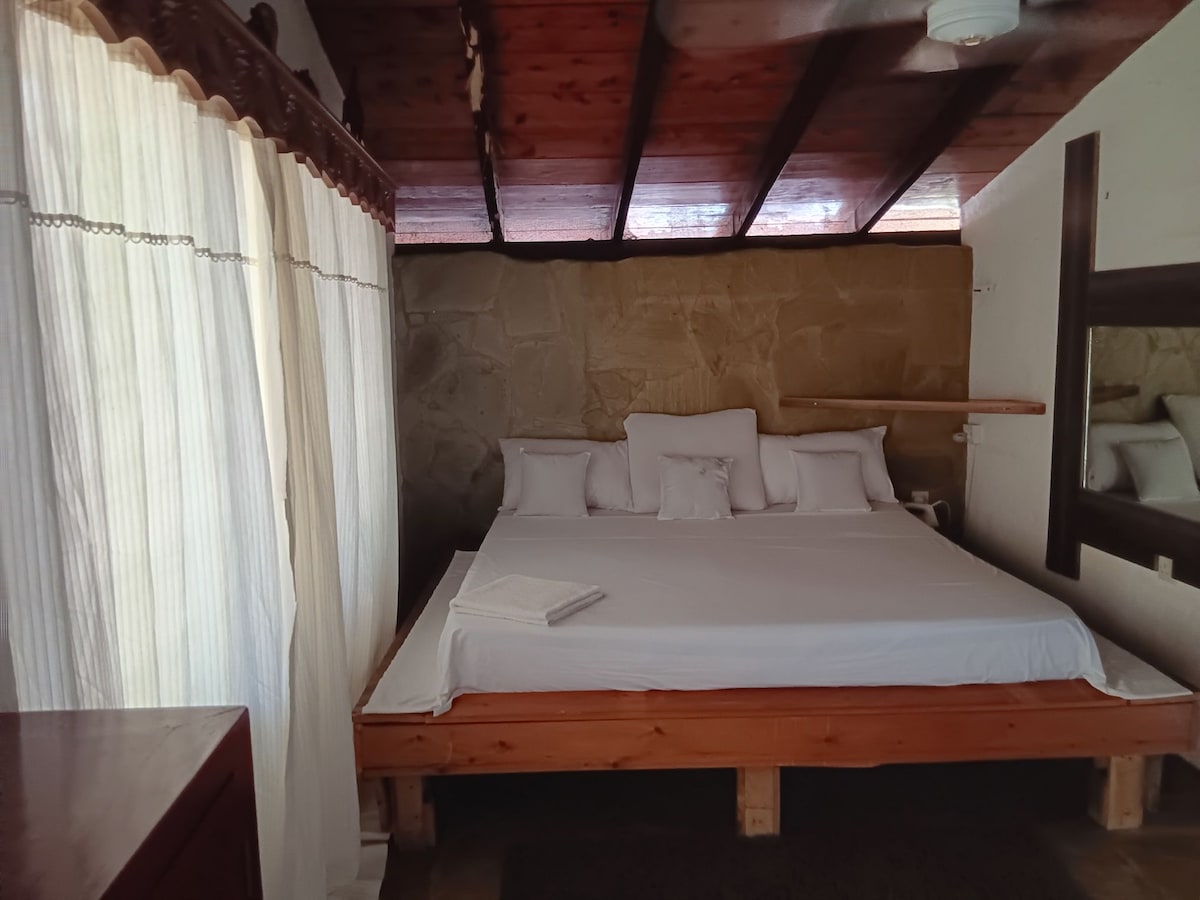 The sleeping area features a spacious bed with multiple white cushions arranged neatly. Natural light filters through a sheer curtain, illuminating the stone wall backdrop. A wooden shelf is positioned above the bed, and a mirror is mounted on a wall, enhancing the room's sense of openness.