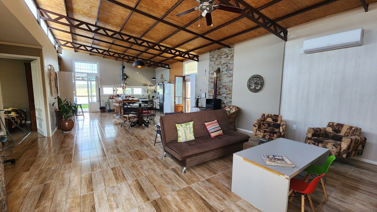 A spacious living area features a comfortable sofa with colorful cushions, a light wooden coffee table, and a dining space with multiple chairs. Natural light floods the room through large windows, highlighting the wood and stone elements in the design.