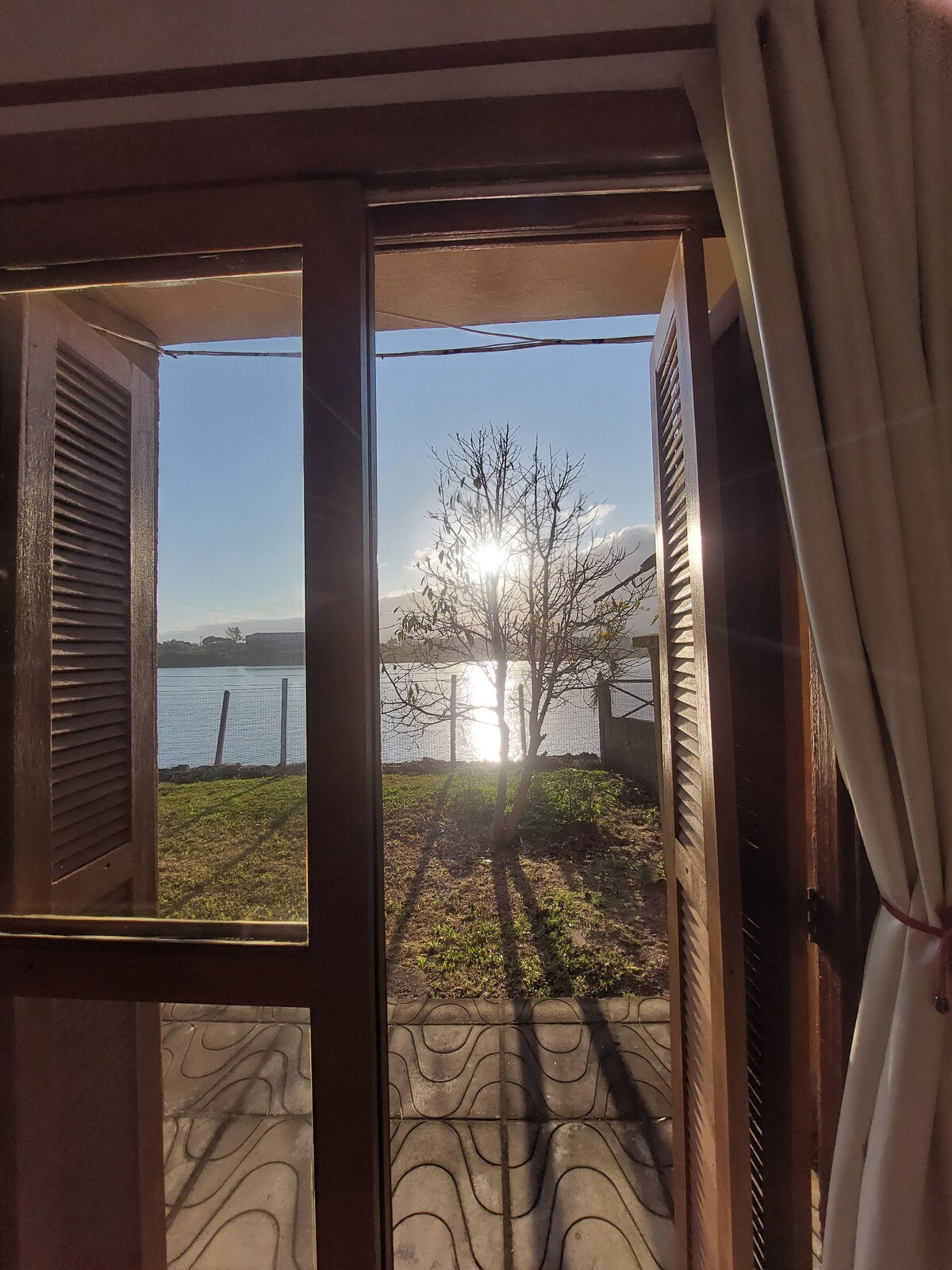 Natural light floods the room through double wooden doors that open to a serene view of the lake. A tree stands near the water's edge, with reflections shimmering on the surface. Gentle shadows from the branches create a peaceful atmosphere.