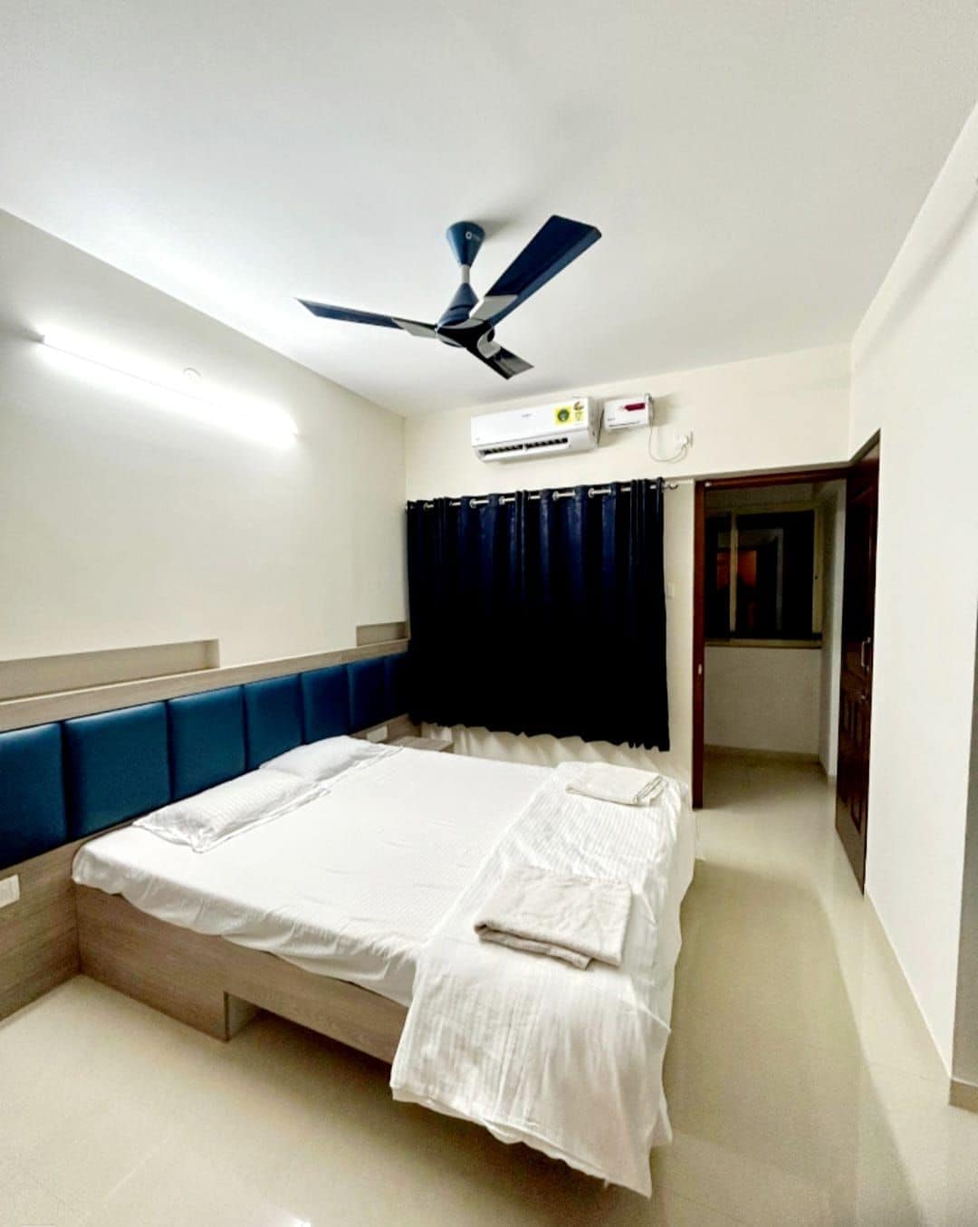 A bright bedroom features a spacious bed with a light-colored bedspread. Dark curtains frame a large window, and a ceiling fan is positioned overhead. The walls are adorned with light shades, creating a calm environment.