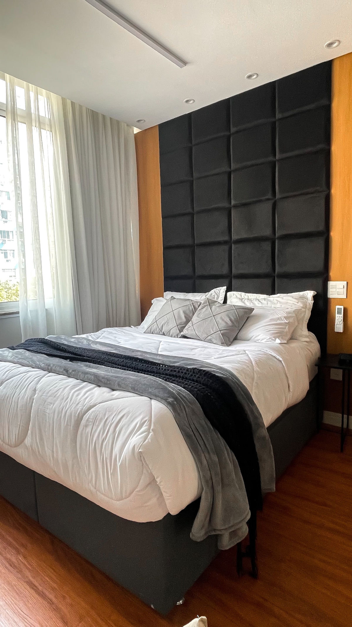 A modern sleeping area features a compact queen bed adorned with layered blankets and decorative pillows. Natural light enters through a large window, complemented by sheer curtains. The headboard displays a textured design, enhancing the room's contemporary feel.