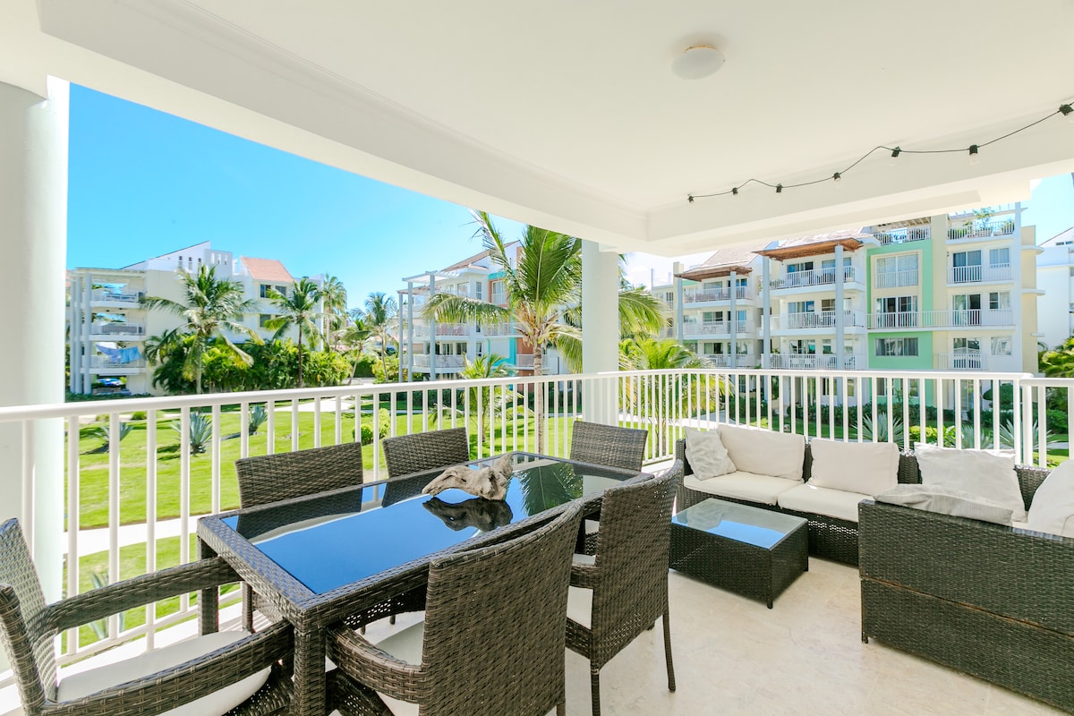 A covered balcony features a large glass-top dining table surrounded by wicker chairs, providing a comfortable seating area. Lush greenery and the exterior of nearby buildings are visible in the background, contributing to a serene outdoor atmosphere.