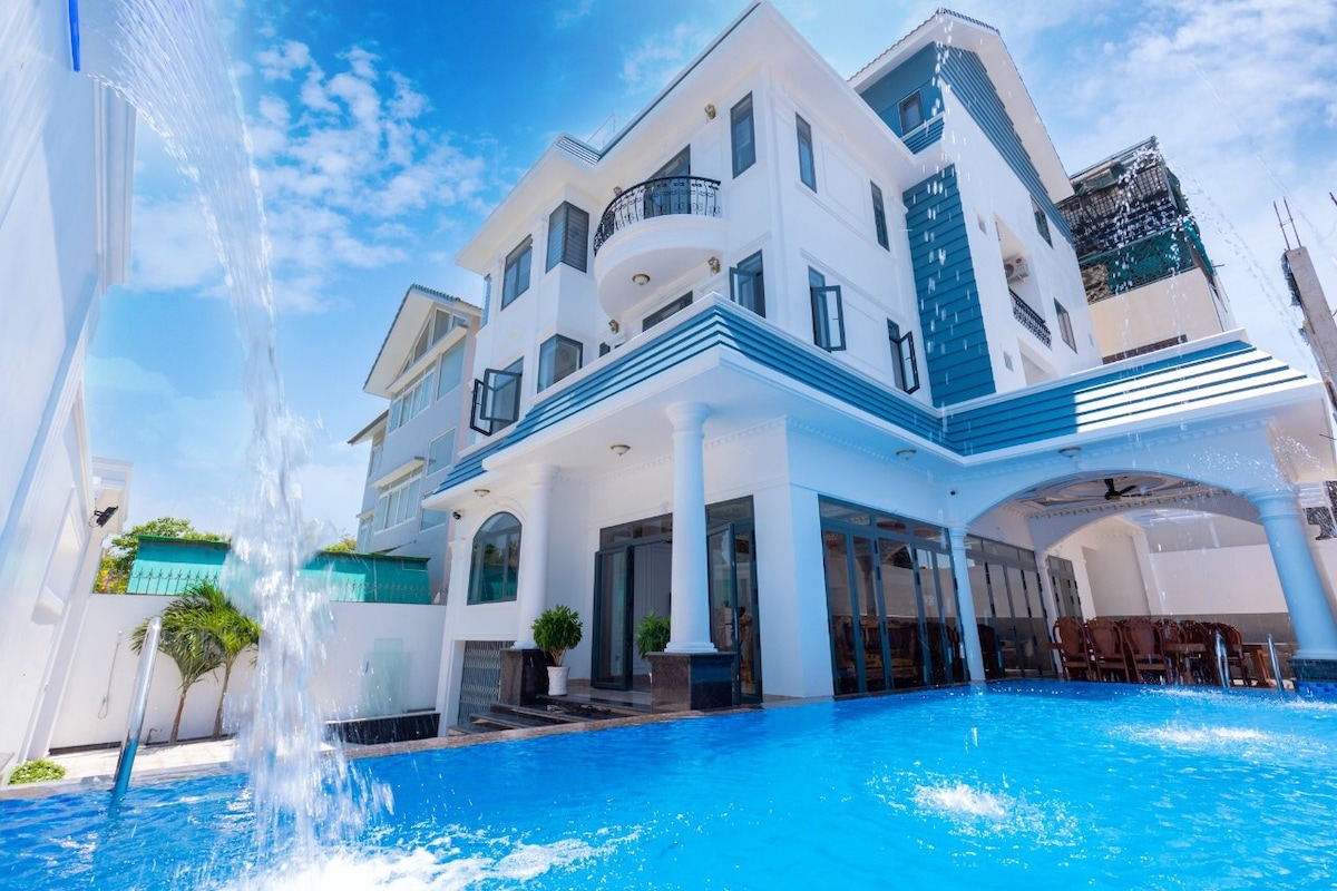 A modern multi-story building is framed by a bright blue sky. The inviting pool layout features a waterfall feature, with lounging chairs arranged beside the water. Large glass doors provide views of the interior spaces, enhancing the airy atmosphere.