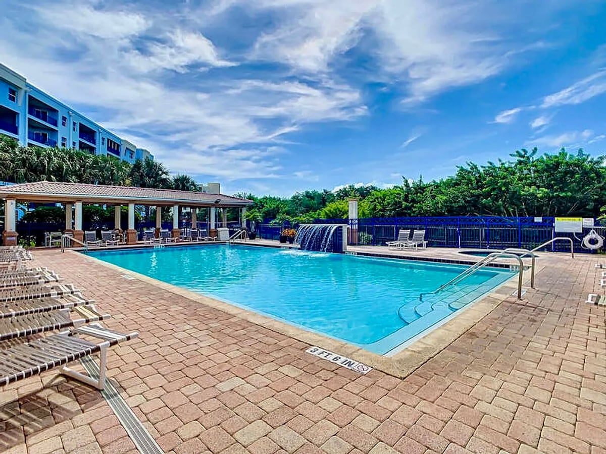 An inviting swimming pool features clear blue water surrounded by a spacious deck. Lounge chairs are positioned along the edge, and a waterfall cascades into the pool area, adding a serene element. Lush greenery is visible in the background, enhancing the tranquil setting.