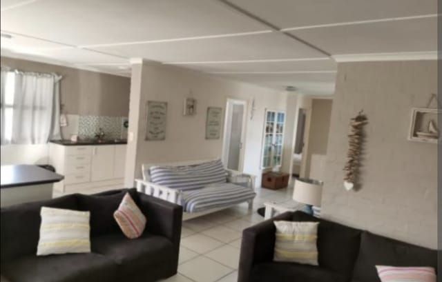 An open living space is showcased, featuring two dark couches with decorative pillows. A striped sofa offers additional seating, while a kitchen area is visible in the background. Natural light enters through a window, illuminating the room's neutral-toned walls and tiled flooring.