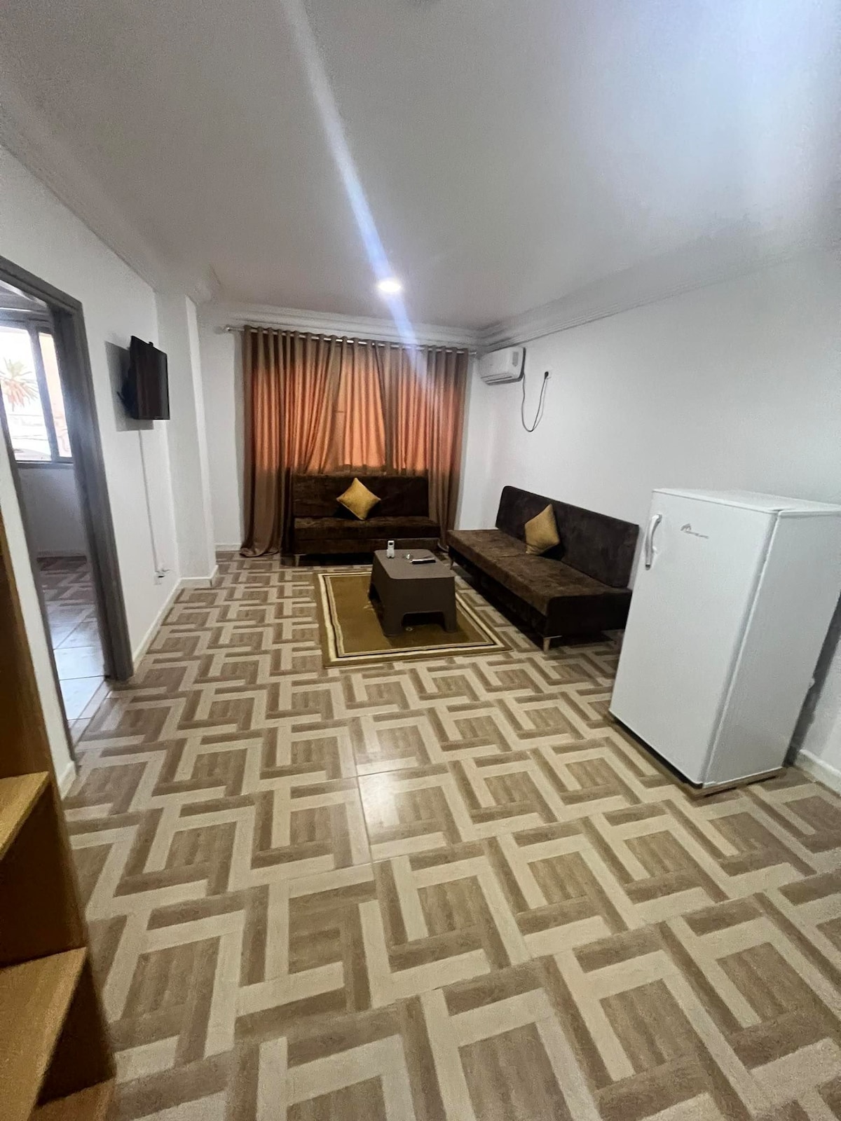 A bright living area features two brown sofas with yellow cushions arranged around a coffee table. A refrigerator stands against one wall, while a wall-mounted television is visible. Natural light enters through a window draped with curtains, creating a cozy atmosphere.