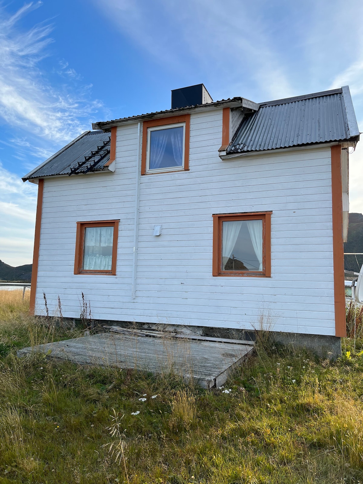 A two-story house is presented from the side, featuring a white exterior with orange trim. Gabled roofs with a mix of metal and shingles cover the structure. Large windows are seen on the upper level, while a wooden platform with grassy surroundings is located at ground level.