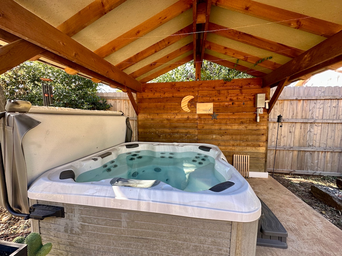A hot tub is situated under a wooden shelter with a sloped roof. The tub features a smooth surface and jets, surrounded by wooden paneling and a gravel floor. Natural light filters in, creating a tranquil space for relaxation.