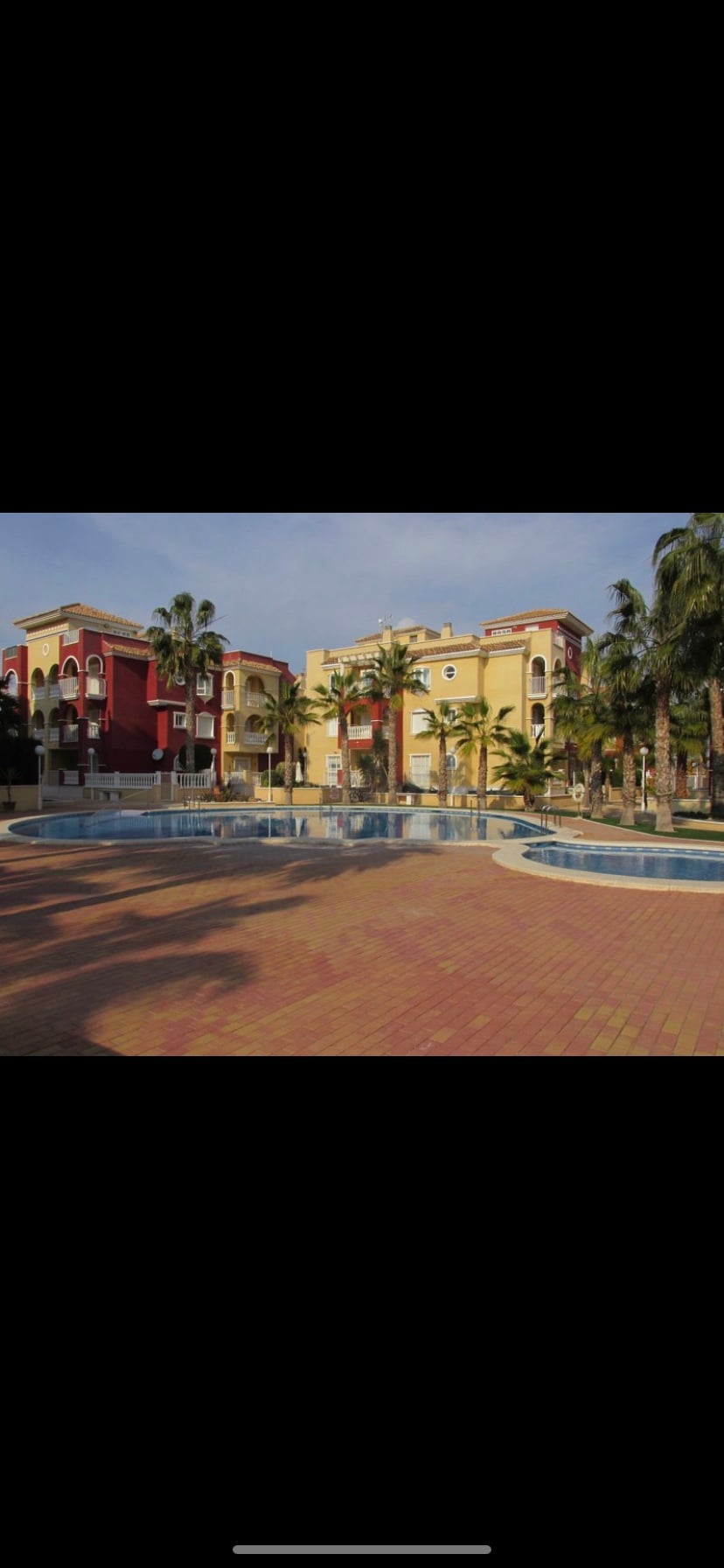 A communal pool area is surrounded by palm trees and colorful buildings in a gated community. Multiple pools are visible, with a spacious deck made of patterned bricks providing ample space for relaxation and leisure. The atmosphere reflects a tranquil environment.