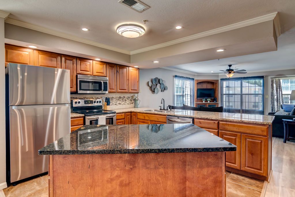 A modern kitchen features dark wood cabinetry and a sizable granite island with seating. Stainless steel appliances, including a refrigerator and stove, are present. Natural light fills the room through large windows, illuminating a comfortable living area in the background.