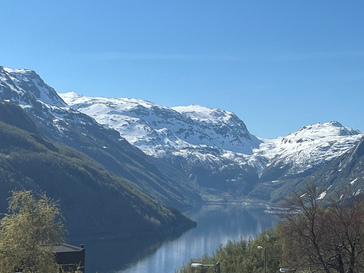 A sweeping view of the mountains and valley is captured, showcasing snow-capped peaks reflected in the calm waters of a lake. Lush greenery lines the foreground, leading to rocky slopes that rise into the distance under a clear blue sky.