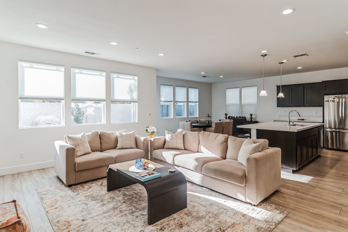 A spacious living area features a light-colored sectional sofa arranged around a dark coffee table. Bright, natural light fills the space through large windows. The adjoining kitchen can be seen in the background, showcasing dark cabinetry and modern appliances.