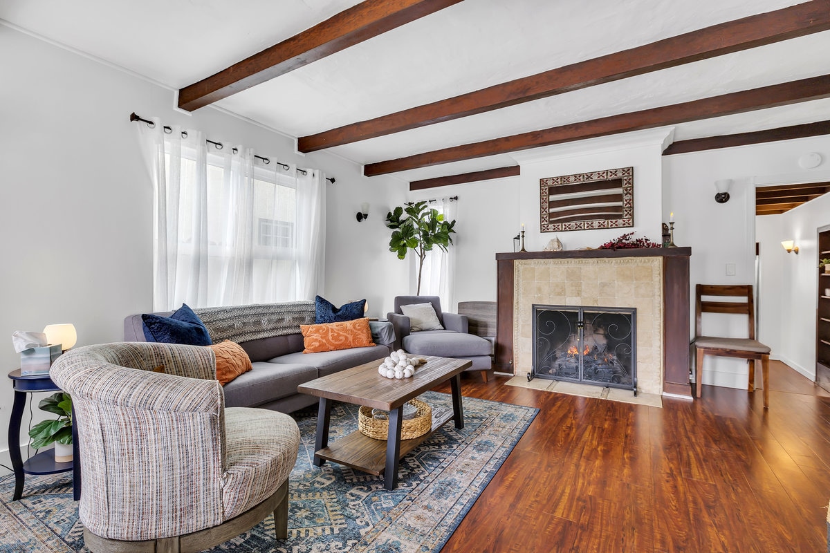 A welcoming living area features a comfortable sectional sofa adorned with decorative pillows. A decorative coffee table rests on a patterned rug in front of a stone fireplace. Natural light filters through sheer curtains, illuminating the rich wood beams overhead.