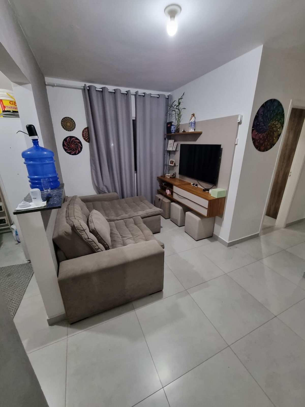 A cozy living area features two light-colored sofas facing a mounted television. A decorative shelf holds personal items, while art pieces adorn the walls. Soft lighting illuminates the space, creating a relaxed atmosphere. Windows are covered by light curtains, allowing for natural light.