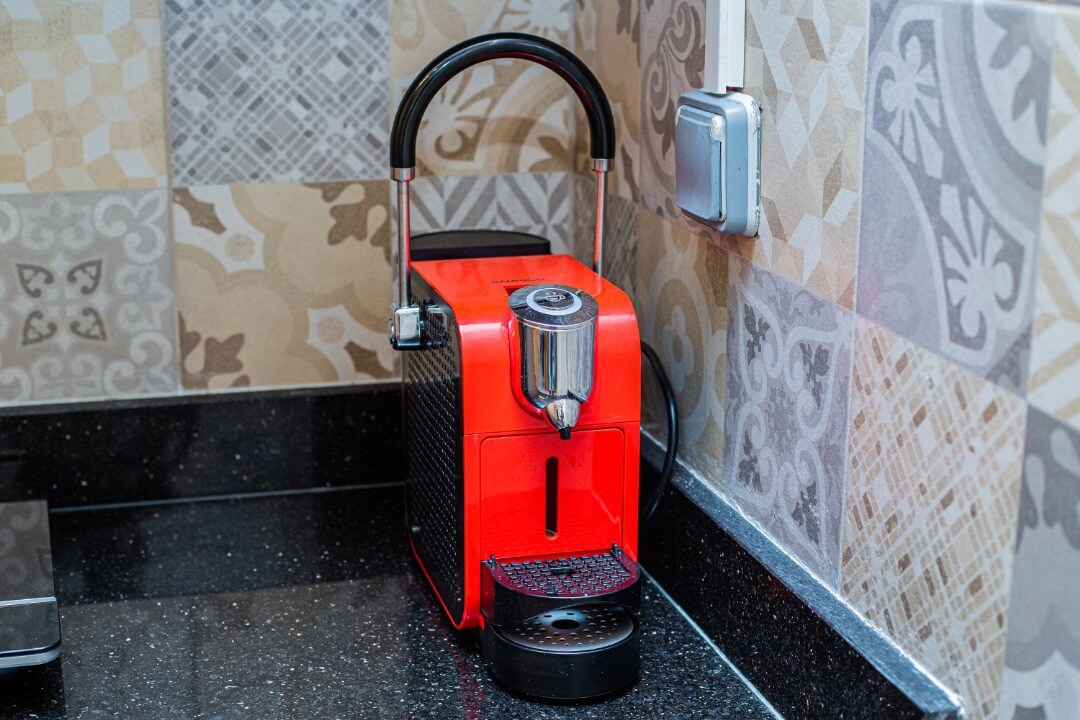 A compact red coffee machine is positioned against a textured wall featuring a mix of patterned tiles. The machine is equipped with a cup holder and a used capsule compartment, complementing the sleek design of the black countertop.