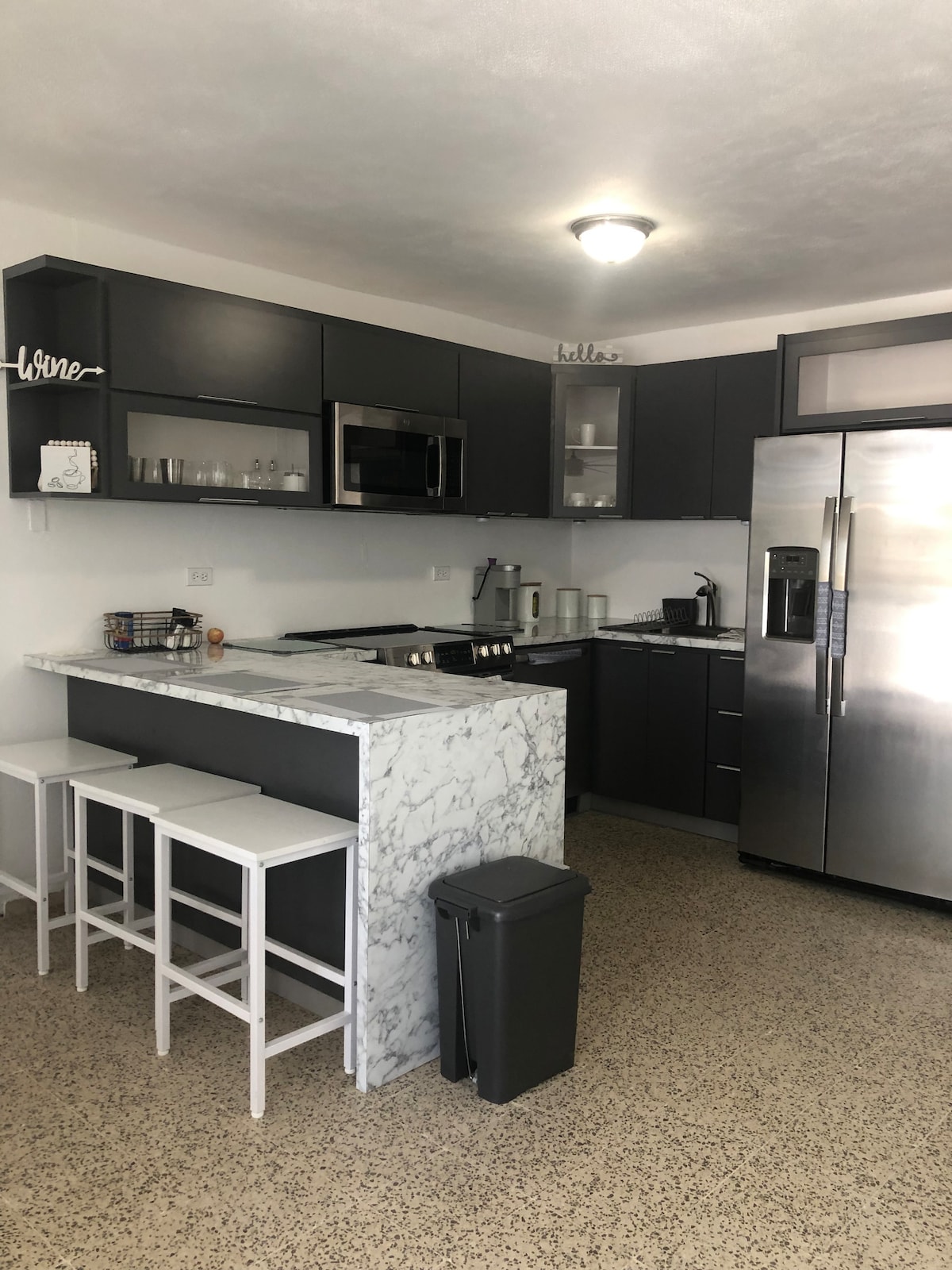 A modern kitchen is equipped with sleek dark cabinetry and stainless steel appliances. A marble-topped breakfast bar with three white stools provides a casual dining space. A large refrigerator and microwave are included, along with various kitchen utensils and dishware visible on the shelves.