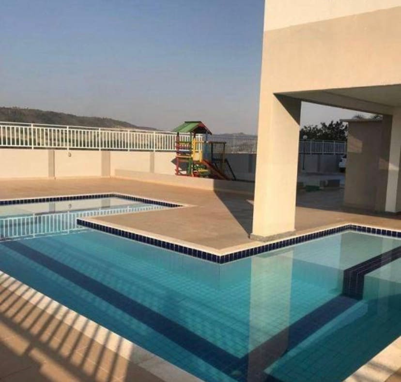 An outdoor pool area features a spacious swimming pool and a separate wading pool, both surrounded by textured tiles. Nearby, a colorful play structure is positioned on a sunlit deck. The backdrop includes distant hills under a clear blue sky.