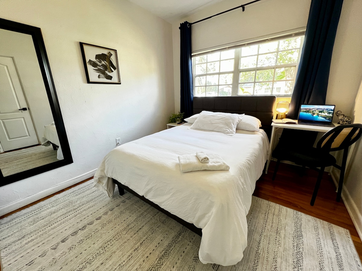 A bright room features a full-sized bed adorned with a white blanket and decorative pillow. A compact desk with a chair is positioned beside a window, which allows natural light to fill the space. A mirror and artwork enhance the minimalist décor on the light-colored walls.