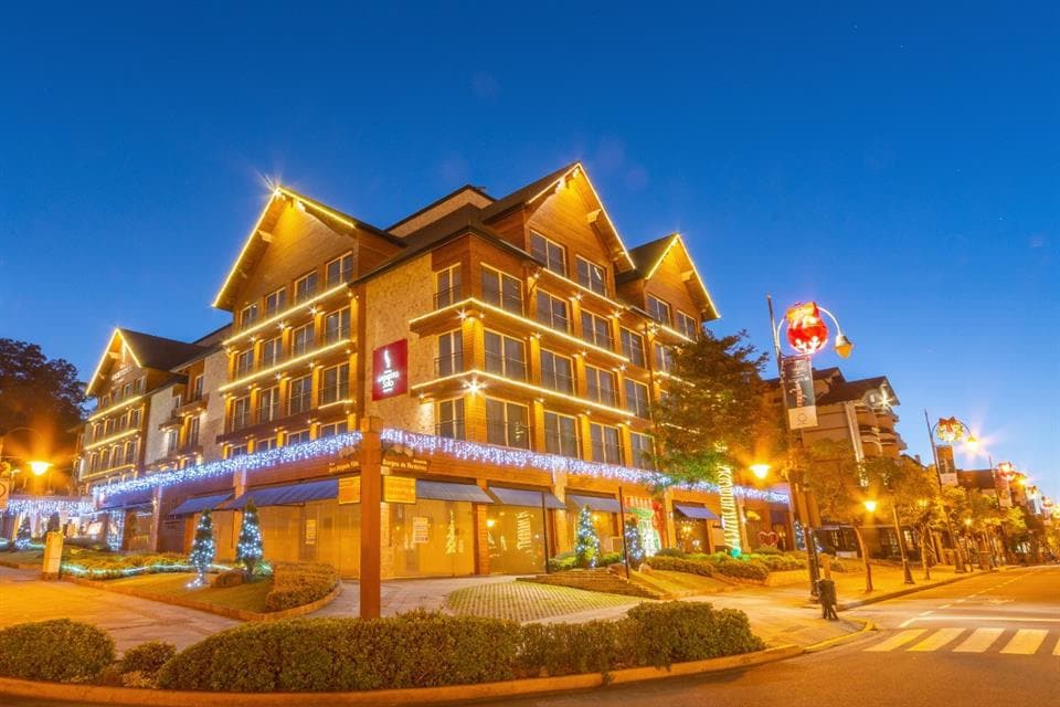 A modern hotel building is illuminated with warm, festive lights against a deep blue twilight sky. Neatly landscaped greenery surrounds the property, while decorative holiday elements enhance the welcoming atmosphere. A tree and light street lamps add to the charm of the exterior.