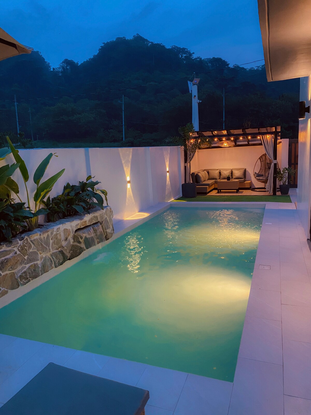 A serene pool area is illuminated by soft lighting, highlighting the clear water. Stone accents line one side of the pool, while lush plants frame the space. A comfortable seating area with a canopy provides a secluded spot for relaxation.