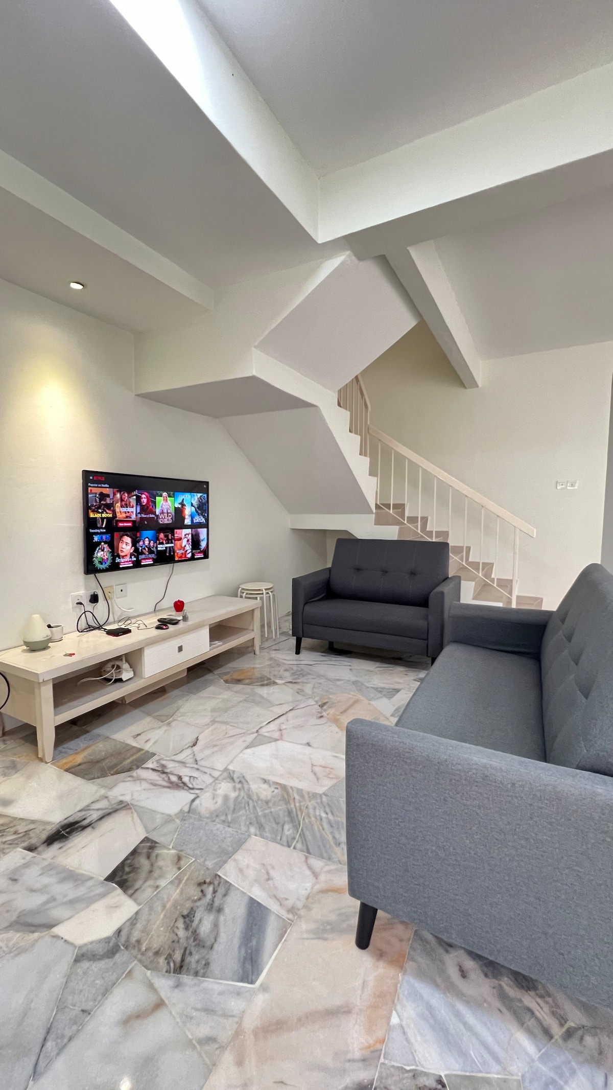 A modern living area features two grey sofas arranged around a light coffee table. A flat-screen television is mounted against a white wall. Marble flooring reflects natural light, while a staircase with a white railing leads to the upper level.