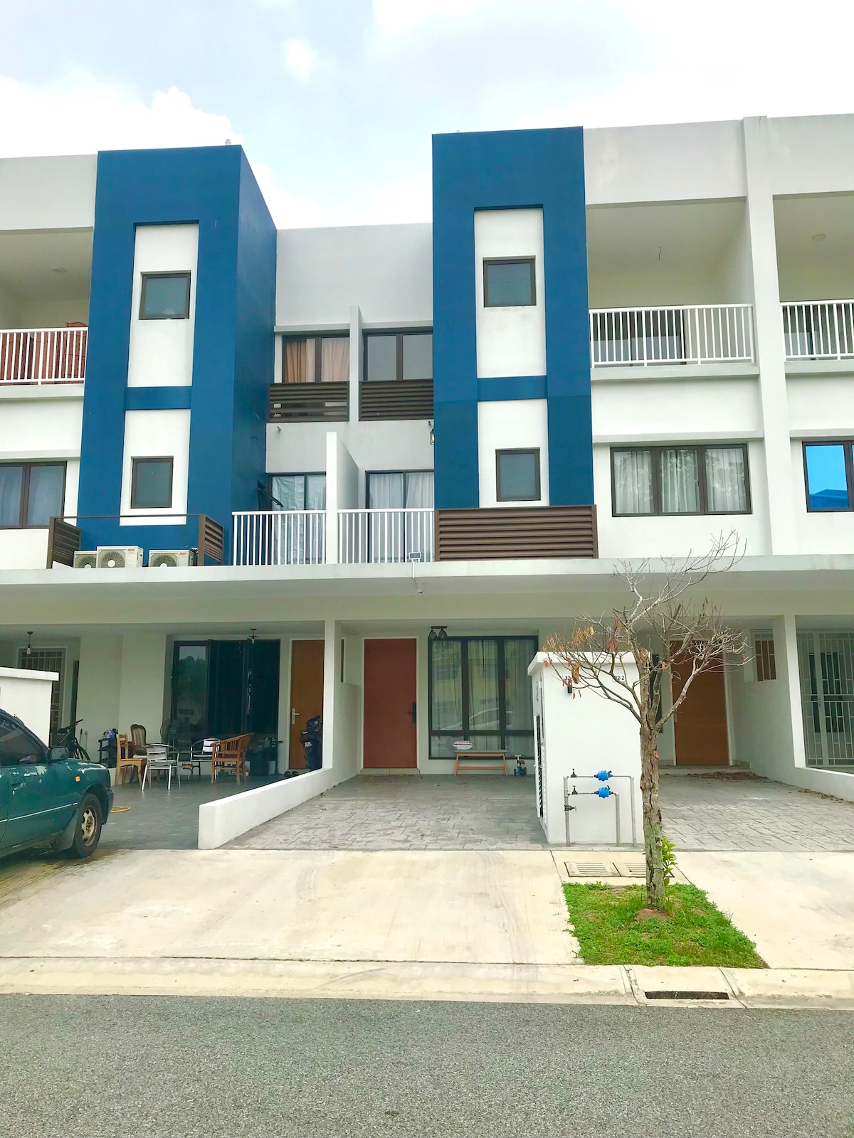 The exterior of a modern townhouse displays a two-story structure with a blue and white facade. Clear balconies are visible on the upper floor, while the ground level features an open porch area and a driveway accommodating two parked vehicles.