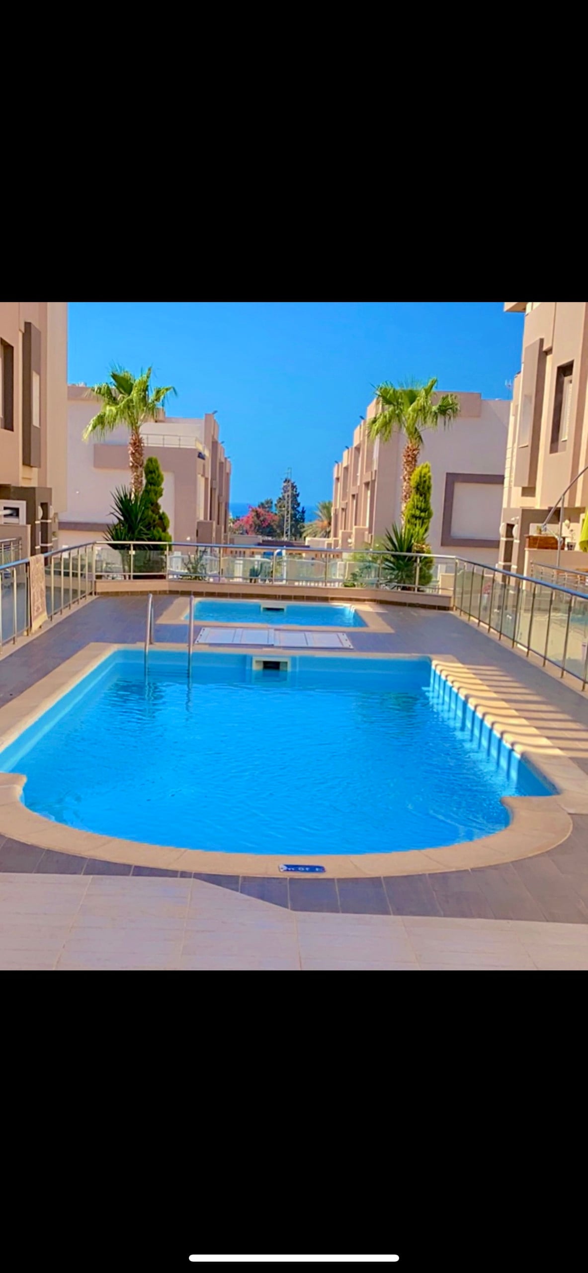 A serene swimming pool is surrounded by palm trees and modern architecture. The pool features a smooth, clear surface reflecting blue skies, with a section for lounging along the edge. Glass railings provide safety and unobstructed views of the inviting water.