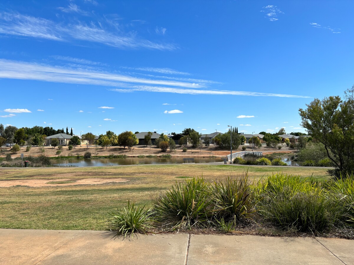 A serene view of a grassy area leading to a calm waterway, framed by lush green plants. Houses are visible in the background, amidst trees under a clear blue sky dotted with wispy clouds.