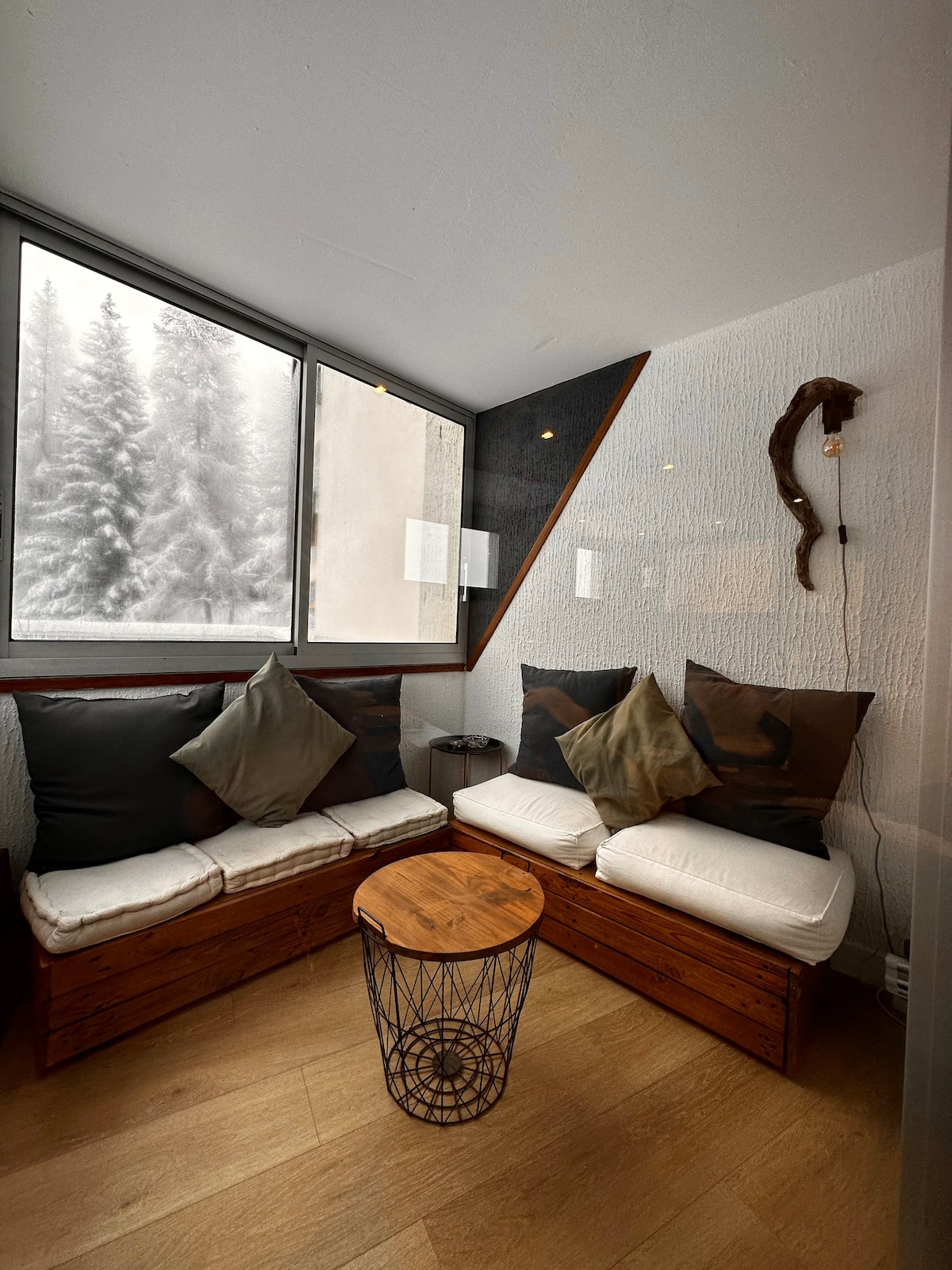 A cozy seating area features a low wooden coffee table surrounded by built-in benches with soft cushions. Large windows provide views of the snowy landscape outside, allowing natural light to illuminate the space.