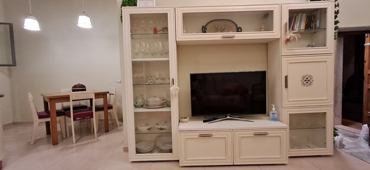 A neutral-toned living area is presented, featuring a large entertainment unit with glass cabinet space displaying dishware. A flat-screen TV is positioned centrally. Behind, a dining table with four chairs offers a cozy space for meals, illuminated by pendant lighting.