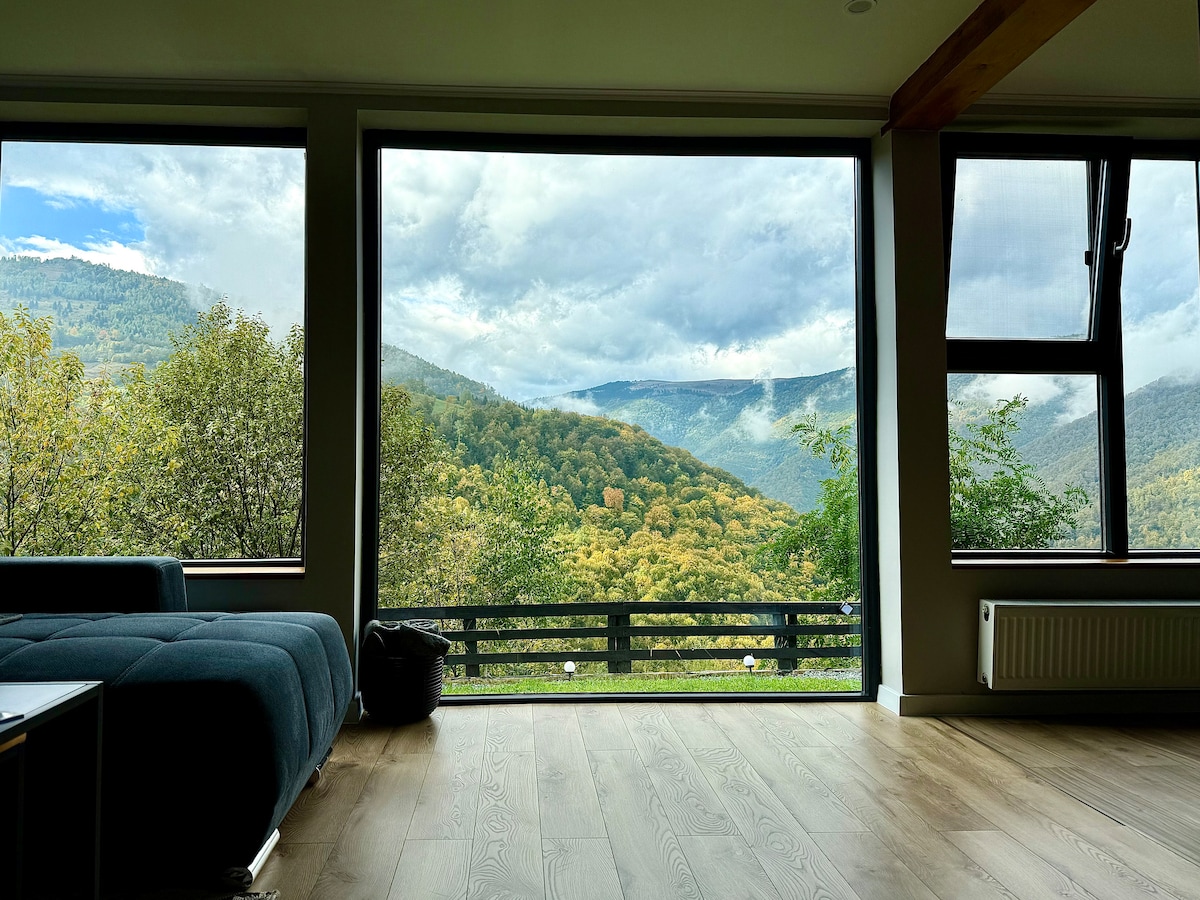Large windows offer panoramic views of the surrounding mountains and valleys, showcasing lush greenery and the changing colors of nature. Natural light floods the spacious living area, highlighting the comfortable seating arrangement and the serene atmosphere.