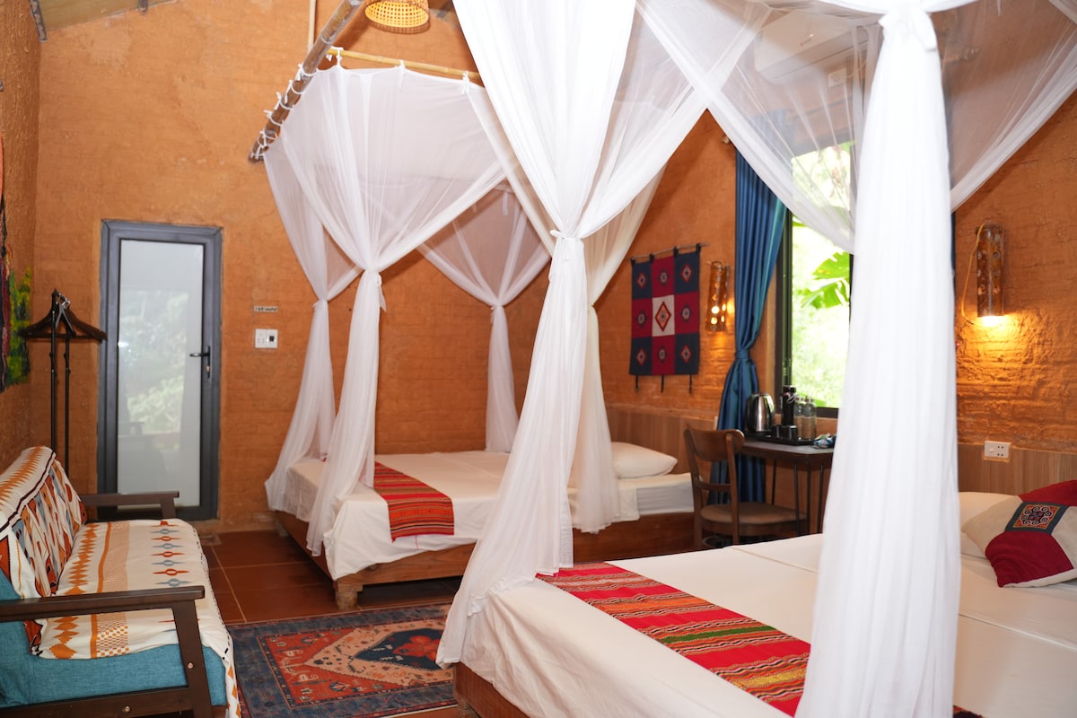The family room features two large queen-size beds draped with white mosquito nets, creating an airy ambiance. A cozy seating area with a wooden bench is present, along with a small dining table. Natural light filters in through the window, enhancing the warm tones of the earthy walls.