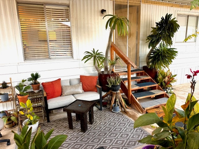An inviting outdoor seating area features a cozy arrangement of potted plants and a patterned rug. A small wooden table is surrounded by comfortable cushions on a sofa. Steps lead up to the entrance of the mobile home, complemented by lush greenery.