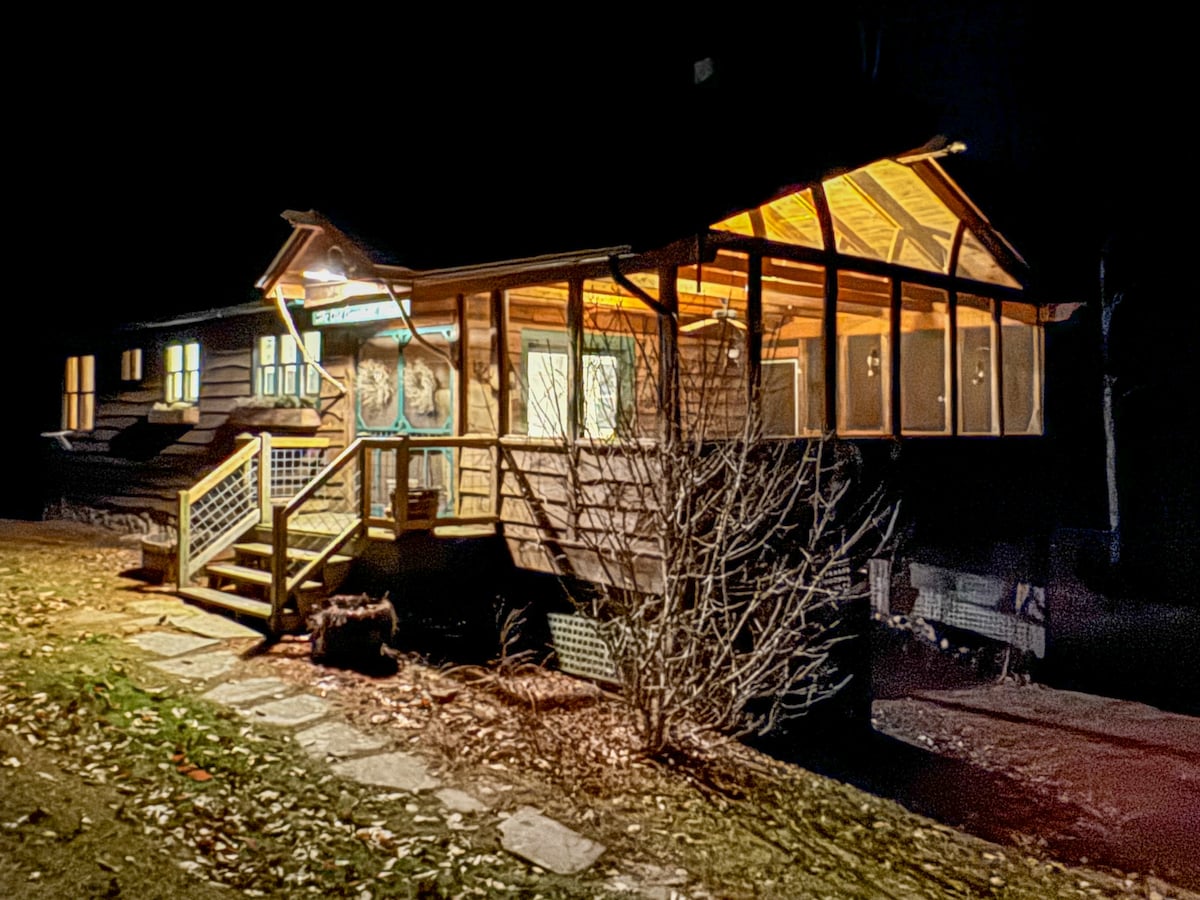 The exterior of a rustic cabin is illuminated softly at night, showcasing a large screened-in porch with wooden railings. Steps lead up to the entrance, and surrounding foliage is visible, creating a serene setting. The structure reflects a welcoming ambiance amidst the quiet landscape.