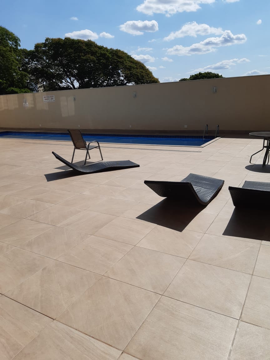 An outdoor area features a swimming pool surrounded by a tiled deck. Two lounge chairs are positioned near the pool, accompanied by a small table nearby. Trees can be seen in the background, and a clear sky is visible above.
