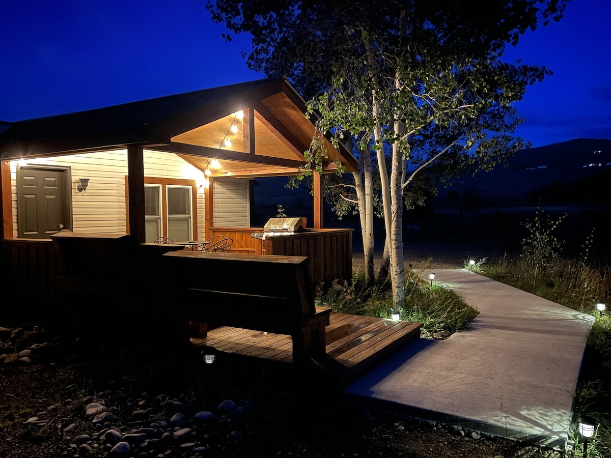 An outdoor view of the porch area, illuminated by soft lights at dusk. A pathway leads to the entrance, flanked by green grass and decorative stones. A grill is positioned on the porch, with comfortable seating visible nearby.