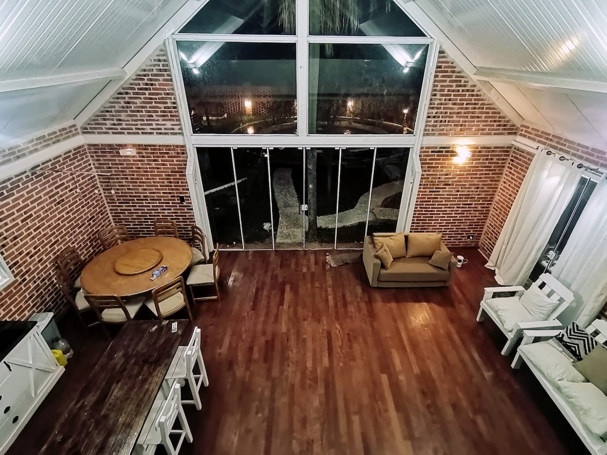 An open living area is showcased with high ceilings and large glass doors that invite natural light. A round dining table sits adjacent to a cozy sofa, while additional seating options include white armchairs. A rich wooden floor complements the brick wall accents.