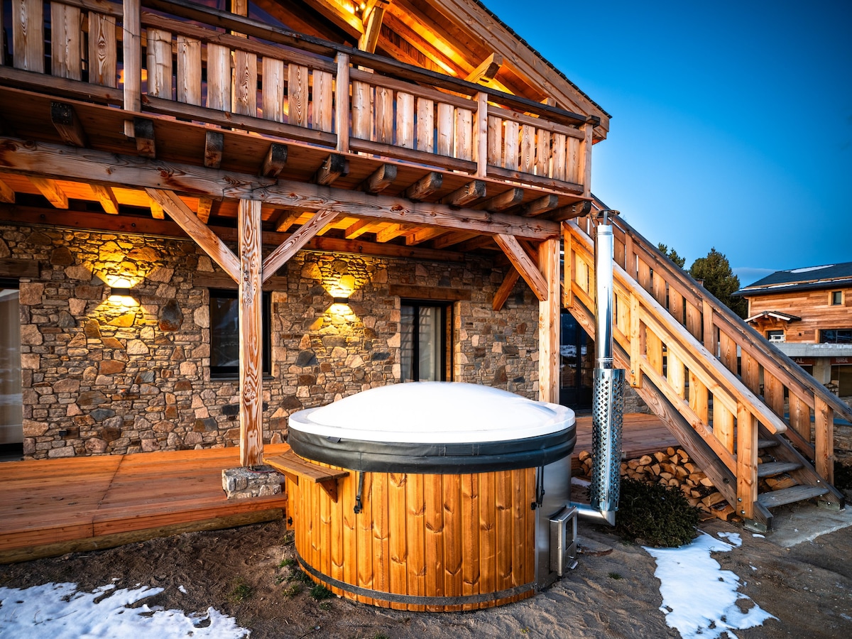 A wooden hot tub, covered with a light layer of snow, is situated beside the stone exterior of the chalet. Warm lighting illuminates the surrounding area, highlighting the textured stone walls and inviting entrance. A wooden staircase leads to the upper level of the chalet.
