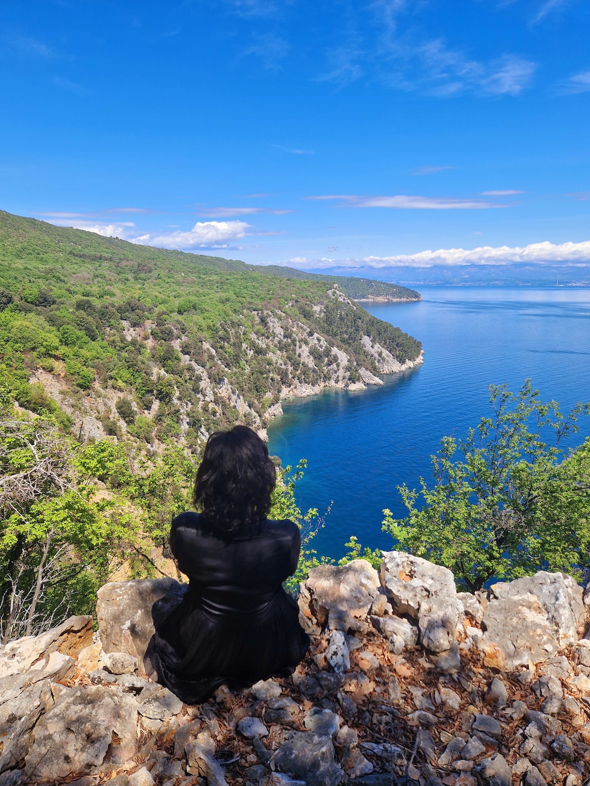 A scenic view of the coastline can be seen, with lush green hills descending into the calm blue waters of Kvarner Bay. The rocky edge in the foreground offers a natural viewpoint, framed by trees and shrubbery, under a clear blue sky.
