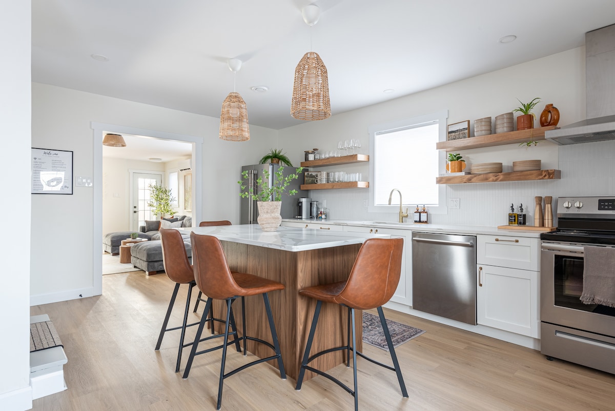 A modern kitchen features a large island with a marble countertop and four leather bar stools. Open shelving displays various dishes and glassware, while stainless steel appliances complement the clean design. Natural light enters through a window, enhancing the inviting atmosphere.