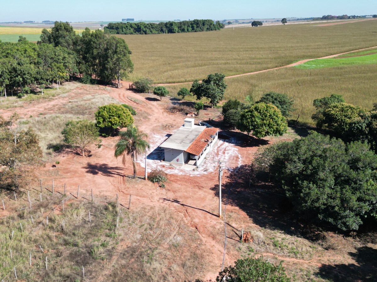 An aerial view showcases a tranquil property surrounded by lush greenery and open fields. The single-story house features a tiled roof and is partially shaded by trees. The spacious outdoor area includes dirt paths and a mix of vegetation, highlighting the property's natural setting.