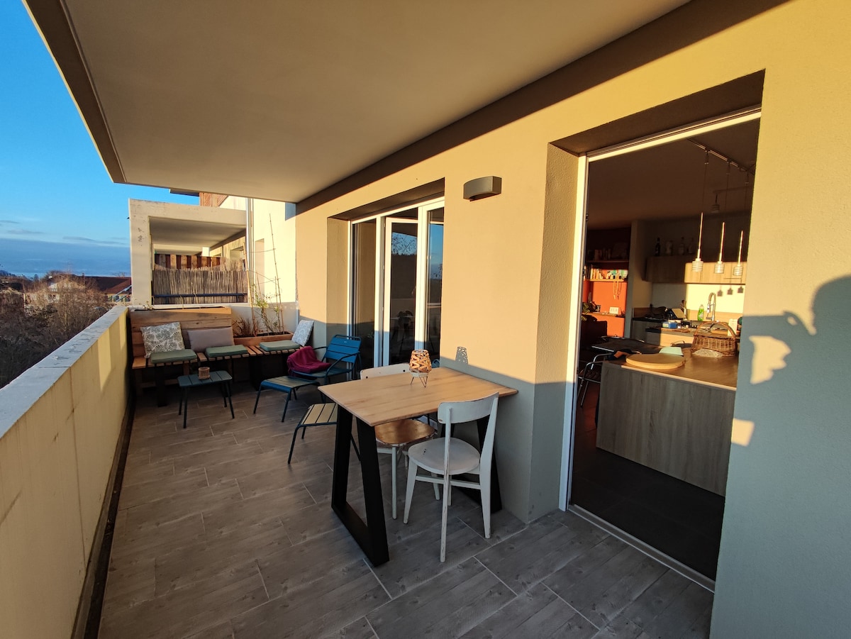 A spacious terrace is viewed, furnished with a wooden dining table and a mix of colored chairs. Natural light fills the area, highlighting the open layout that connects to the kitchen. The surrounding view includes distant hills and nearby buildings.