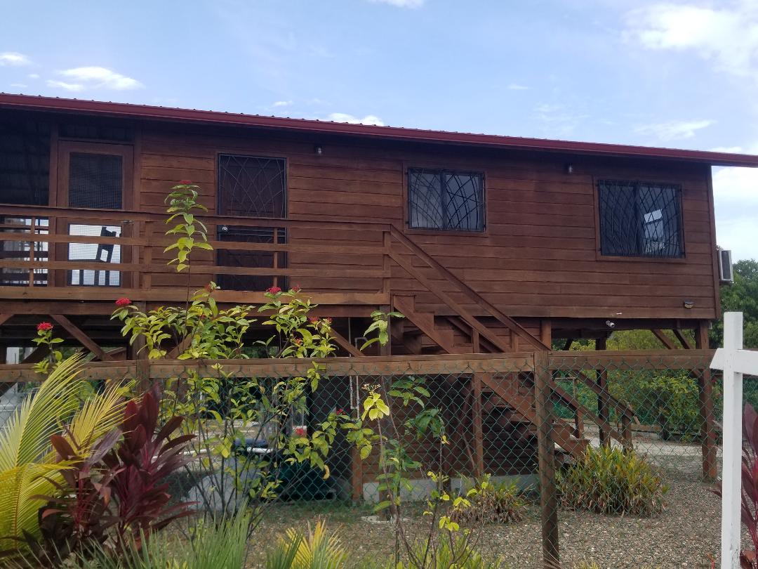 A wooden two-story house stands elevated on stilts, featuring a red metal roof. Dark grid-patterned windows offer a view of the surroundings. A wooden deck extends from the front, providing access to the main entrance and outdoor space, with lush greenery visible in the foreground.