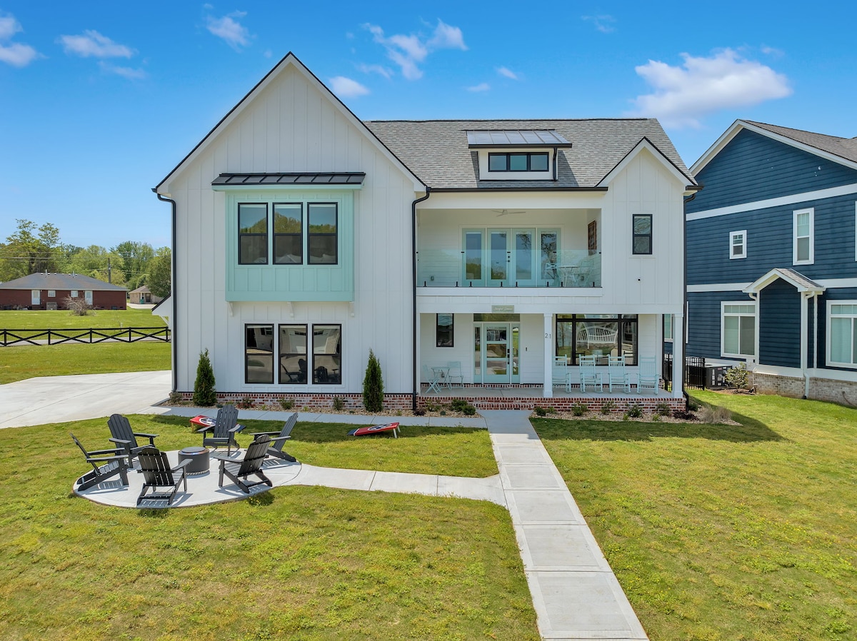 The exterior of a modern two-story house is showcased, featuring a white facade with large windows. A covered balcony is visible on the upper level, complemented by a welcoming front porch below. A circular seating area with Adirondack chairs is positioned in the grassy yard.