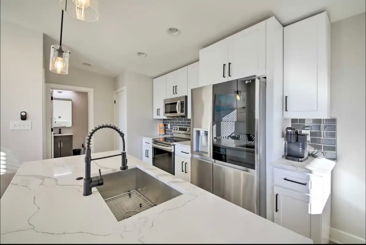 The well-equipped kitchen features modern appliances, including a sleek refrigerator and microwave. A spacious island with a farmhouse sink is complemented by light cabinetry and stylish backsplash tiles. Natural light illuminates the workspace, creating a bright cooking environment.