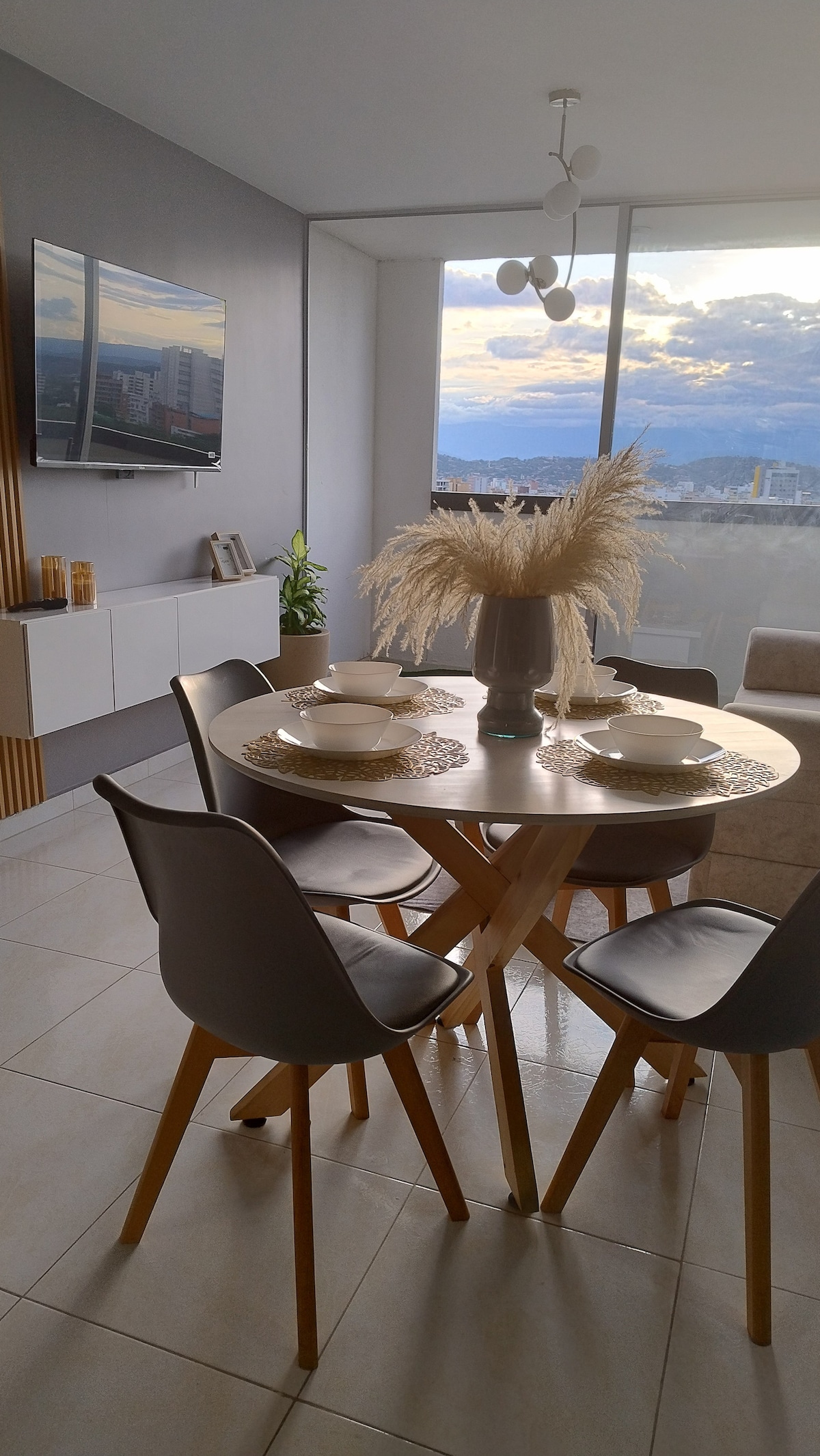 A modern dining area features a round wooden table set for four, accompanied by simple, stylish chairs. A vase filled with dried grass serves as a centerpiece. A flat-screen television is mounted on the wall, while large windows provide a view of the city skyline.