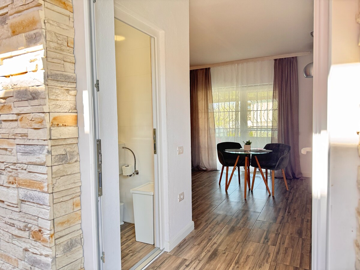 A welcoming entrance leads to a bright living area, featuring two dark chairs and a small table set on light wood flooring. Natural light filters through sheer curtains, enhancing the warm ambiance created by the walls and decor.